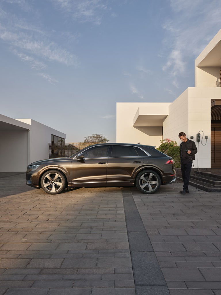 Ein Mann mit Ladekabel steht neben einem braunen Audi Q8 SUV TFSI e vor einem modernen Haus.
