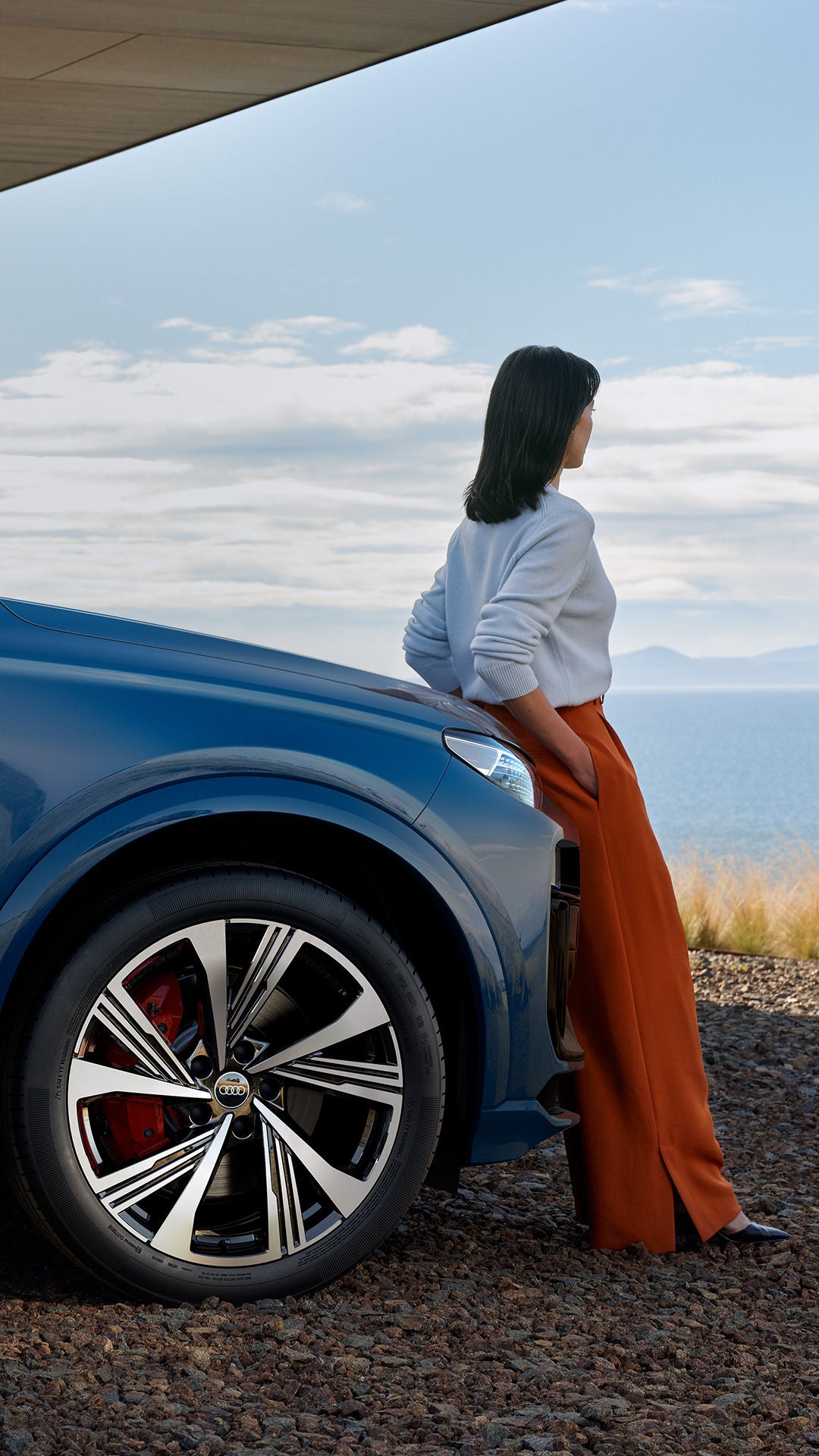 Frau lehnt an einem blauen Audi SUV mit Blick aufs Wasser, im Hintergrund Berge und blauer Himmel mit Wolken.