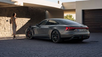 Eine Frau lädt einen grauen Audi RS e-tron GT in einer modernen Einfahrt mit Steinwand und Garage.