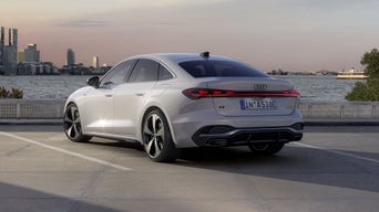 Die A5 Limousine e-hybrid steht im schrägen Heckprofil auf einem Parkplatz am Ufer eines Sees. Dahinter ist die Skyline einer Stadt bei Dämmerung zu sehen.