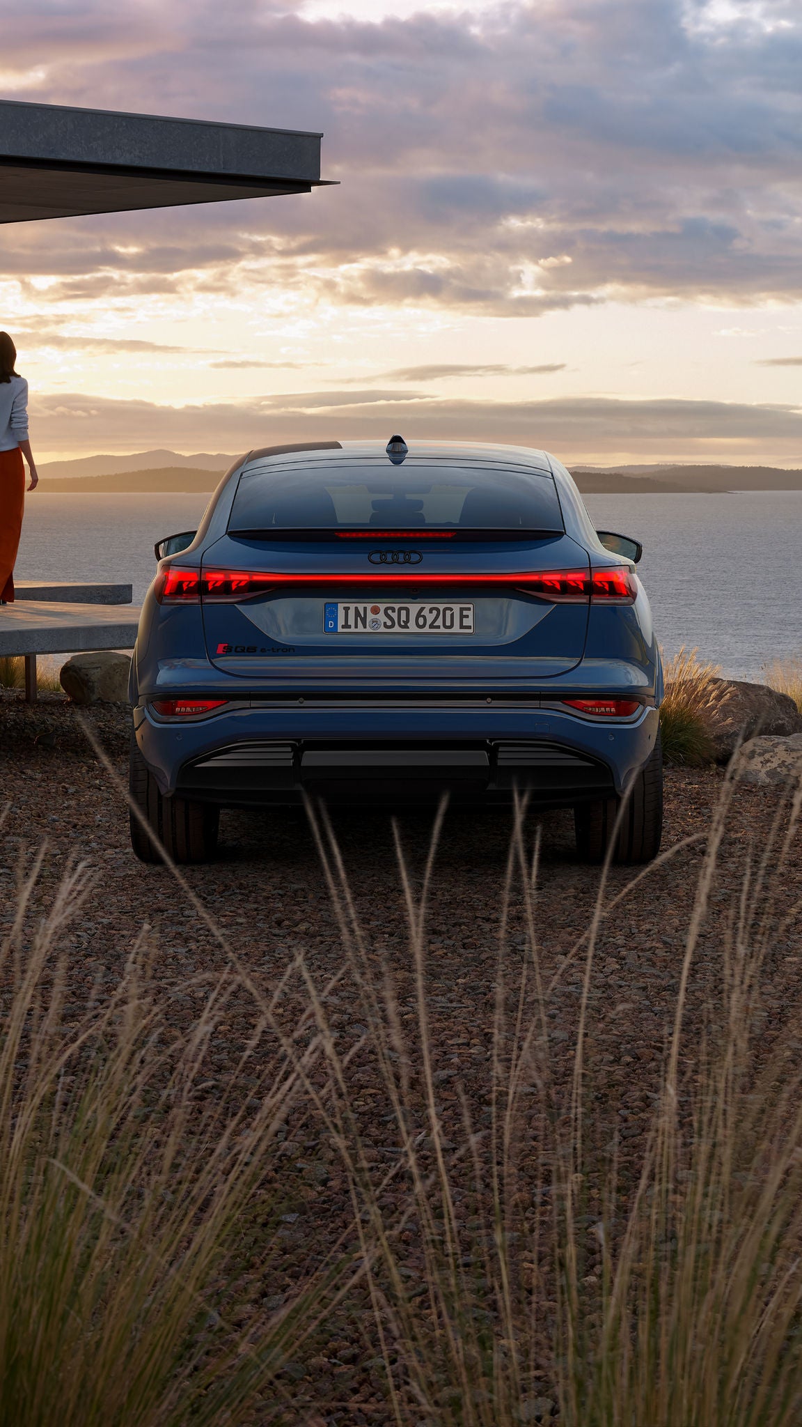 Heckprofil des SQ6 Sportback e-tron, welcher vor einem modernen Haus direkt an der Küste steht. Auf der Veranda steht eine junge Frau und schaut in Richtung des Wassers.