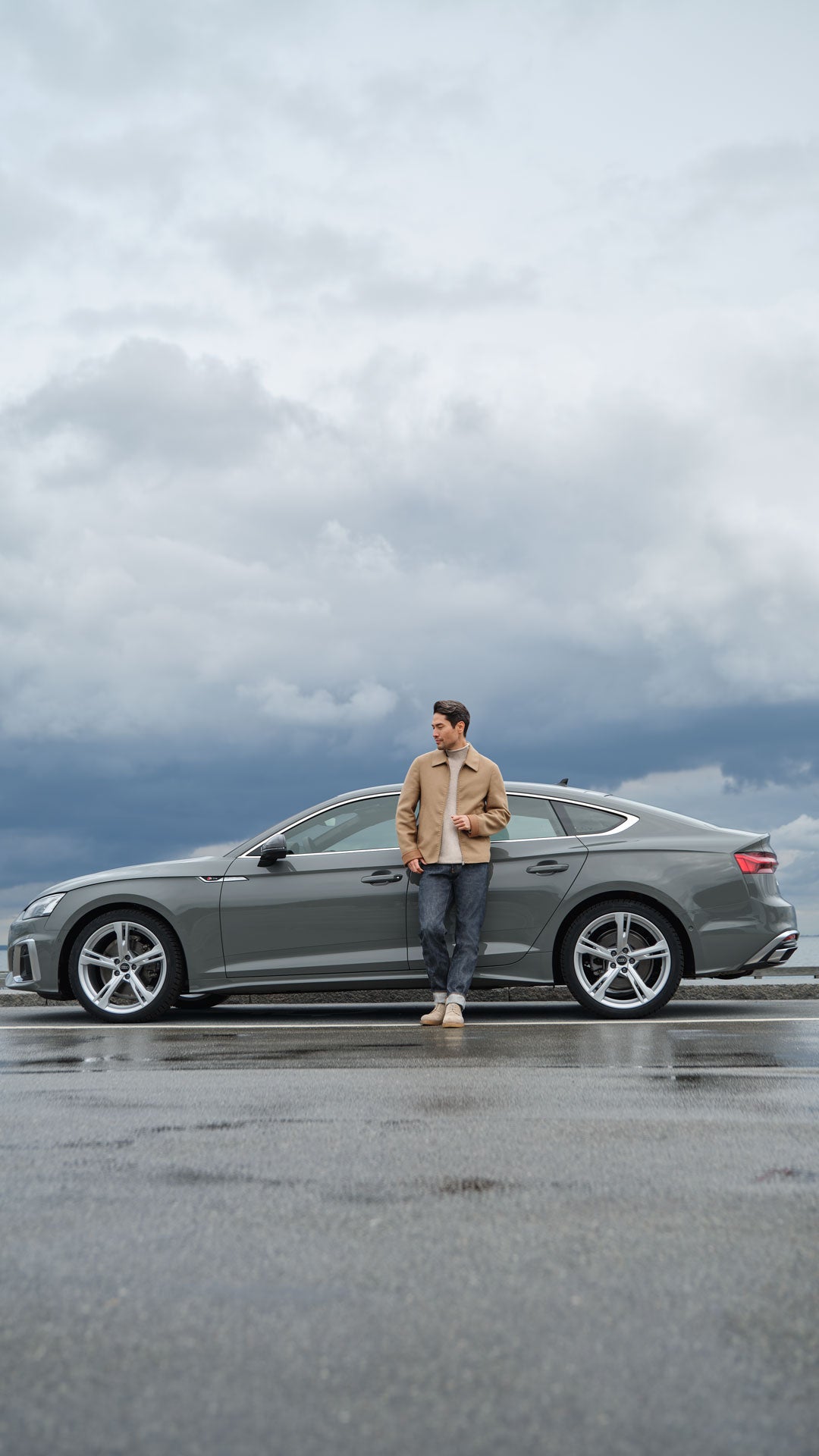 Ein Mann lehnt mit einem Getränk an einem grauen Auto, bei bewölktem Himmel auf nasser Straße nahe dem Wasser.