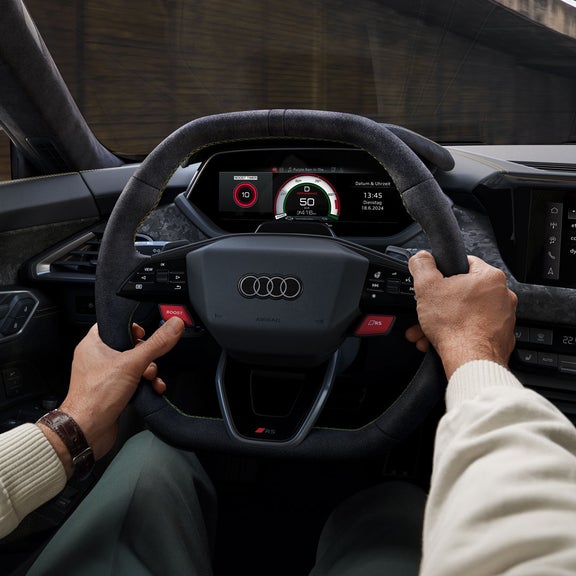 Zwei männliche Hände umfassen das Lenkrad des Audi RS e-tron GT performance. Der linke Daumen betätigt den roten Boost-Button.