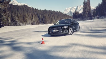 Ein schwarzer Audi A8 50 TDI quattro fährt auf schneebedeckter Strecke durch einen Slalom-Parcours mit Hütchen, umgeben von verschneiten Bergen und Tannen.