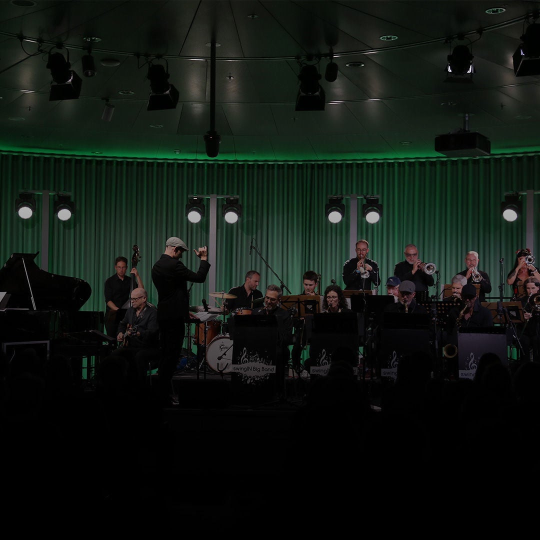Big Band auf Bühne mit Dirigent; Musiker spielen vor grün beleuchtetem Vorhang.