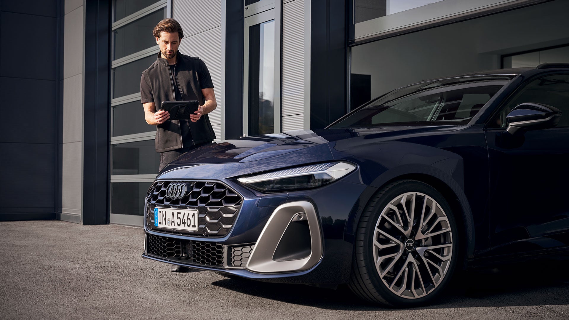 Ein Mann vom Kundendienst steht mit einem Tablet neben einem blauen Audi, der zur Hälfte in einer Toreinfahrt zu einem Gebäude steht.