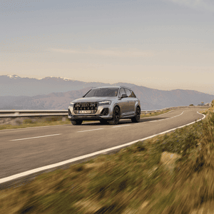 A silver Audi SUV driving on a scenic mountain road under a clear sky, with snow-capped peaks in the distance. The mood is adventurous and serene.