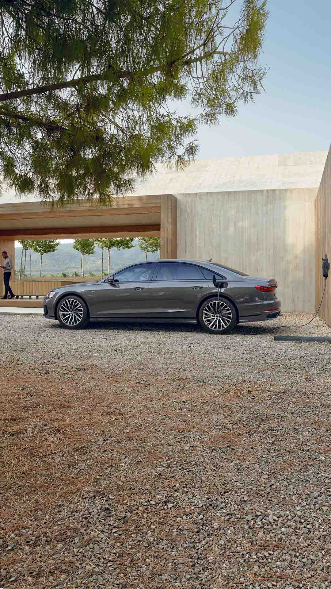 Ein grauer Audi A8 L 60 TFSI e lädt an einer Wandladestation in naturnaher Umgebung mit hellem Holzgebäude. Weit im Hintergrund steht ein farbiger Mann.