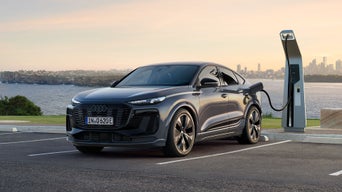Der Q6 Sportback e-tron steht auf einem Parkplatz und ist an eine Ladestation angeschlossen. Im Vordergrund steht ein Mann mittleren Alters und im Hintergrund sind das Meer, sowie die Skyline einer Stadt zu sehen.&nbsp;