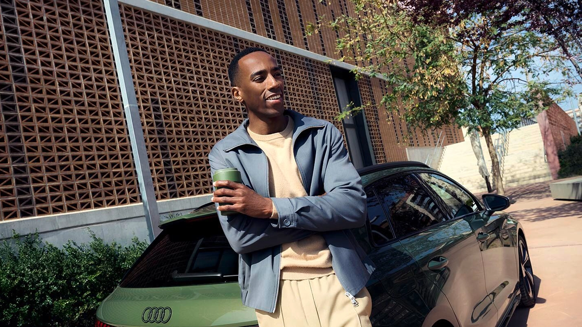 Man in a blue jacket leaning on Audi A3 Sportback, holding green cup.