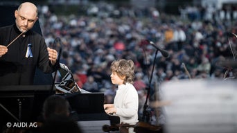Junge spielt am Klavier. Links neben ihm steht ein Dirigent.