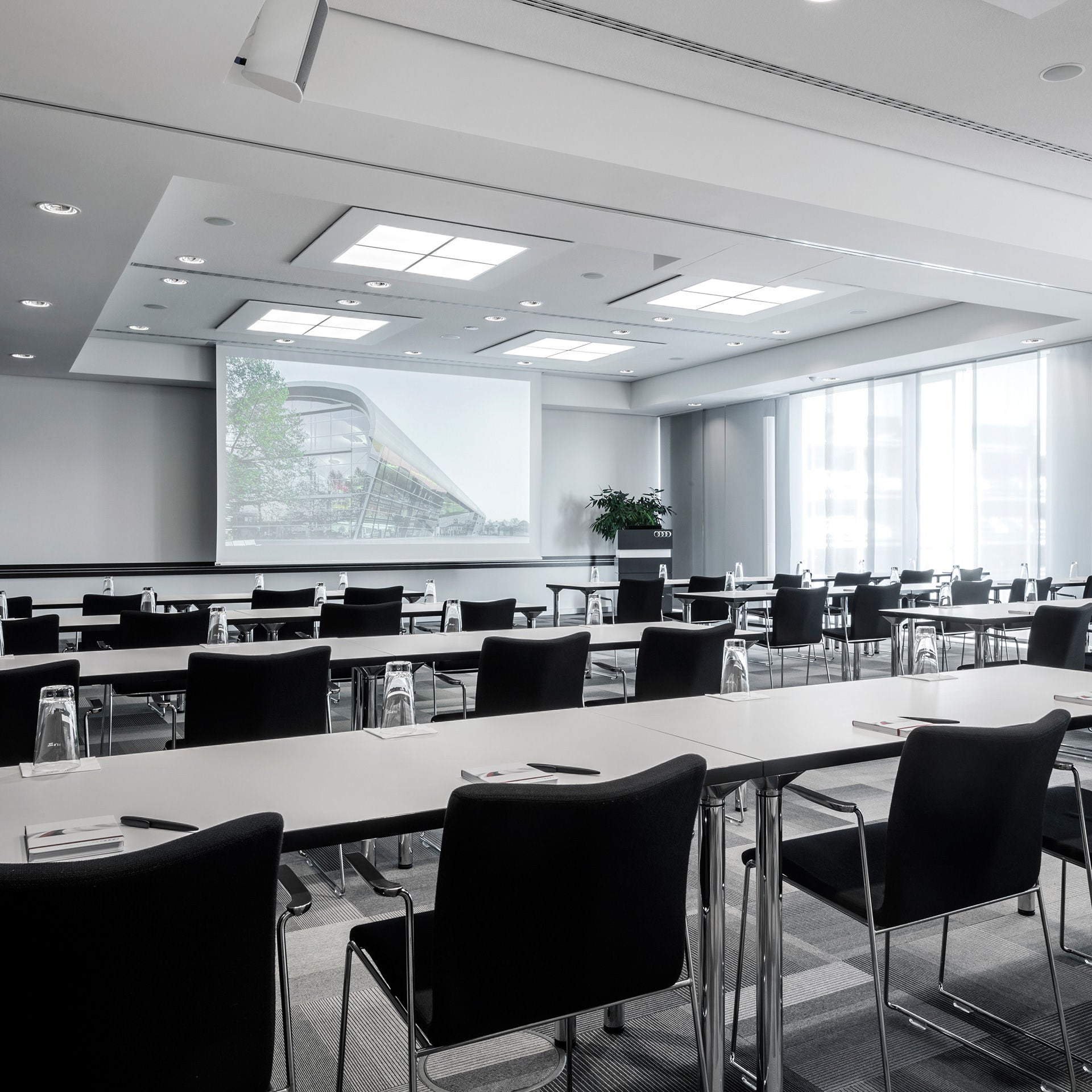 Heller Konferenzraum mit langen Tischen, schwarzen Stühlen, Wassergläsern und einer Präsentation auf der Leinwand.   