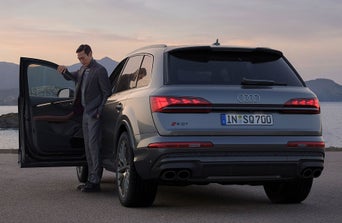 A stylish Audi parked by the water at sunset, with a man in a suit standing next to it. Scenic mountains in the background.