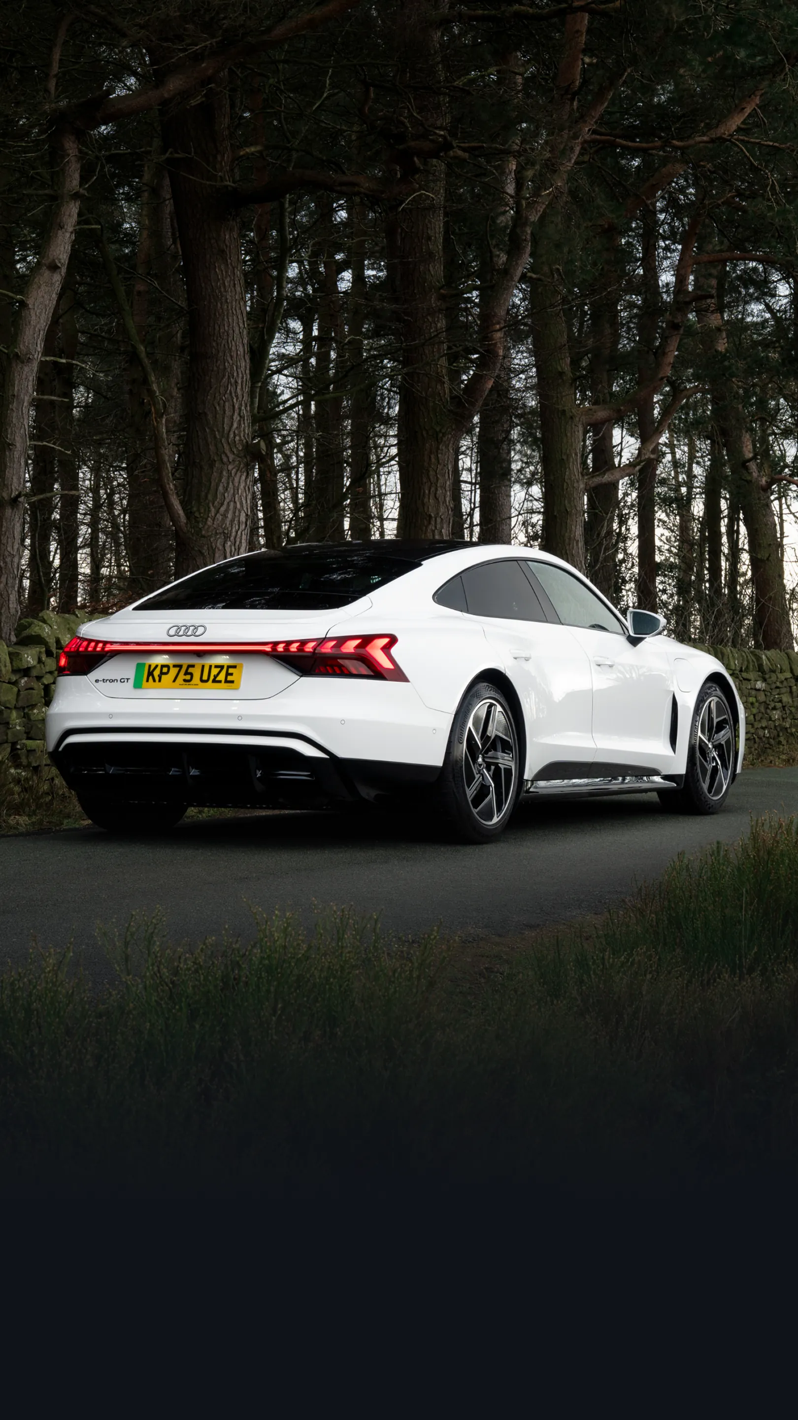 White Audi e-tron GT parked on a quiet country road, shown from the rear three-quarter angle. The vehicle features a sleek fastback silhouette, large aerodynamic alloy wheels and a full-width rear LED light bar. A stone wall and tall pine trees line the roadside, creating a natural woodland backdrop that highlights the vehicle’s sporty design.