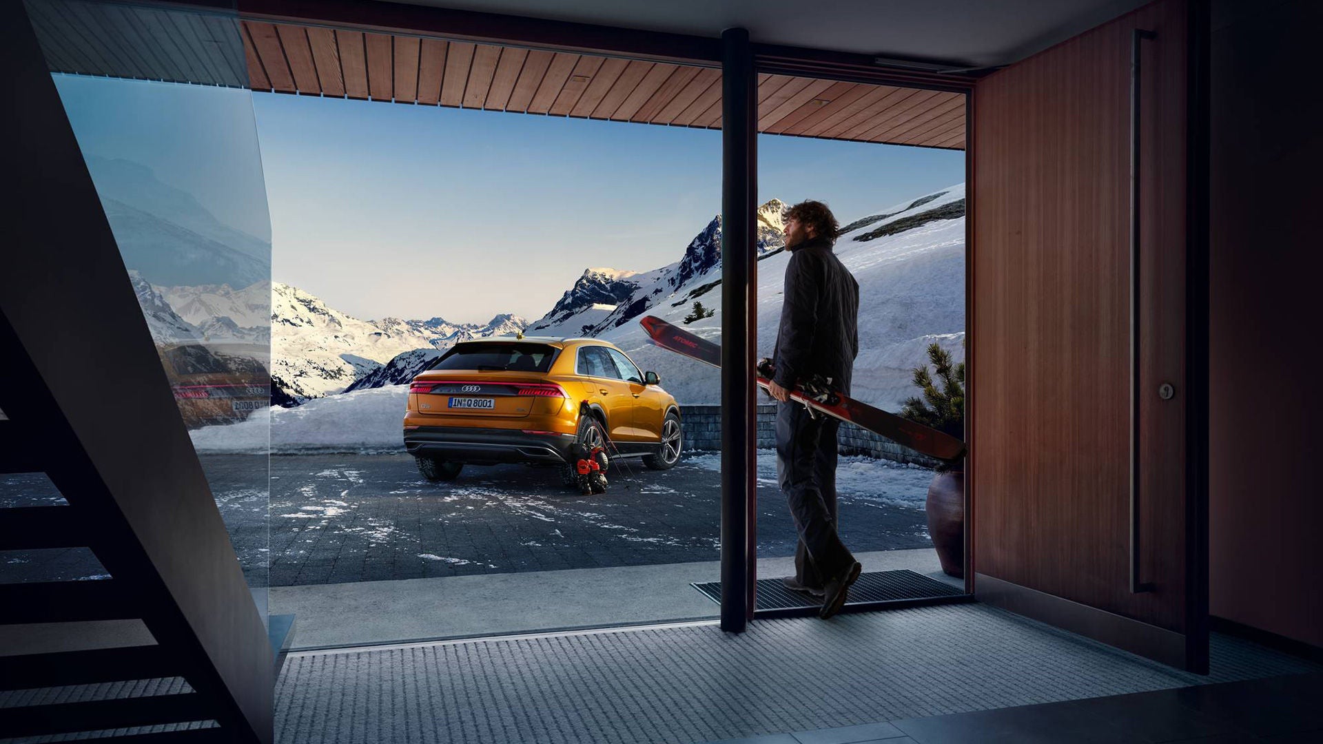 A man looks out of the panoramic window of a modern ski lodge at a snow-covered mountain backdrop and a yellow Audi parked in front of the building.
