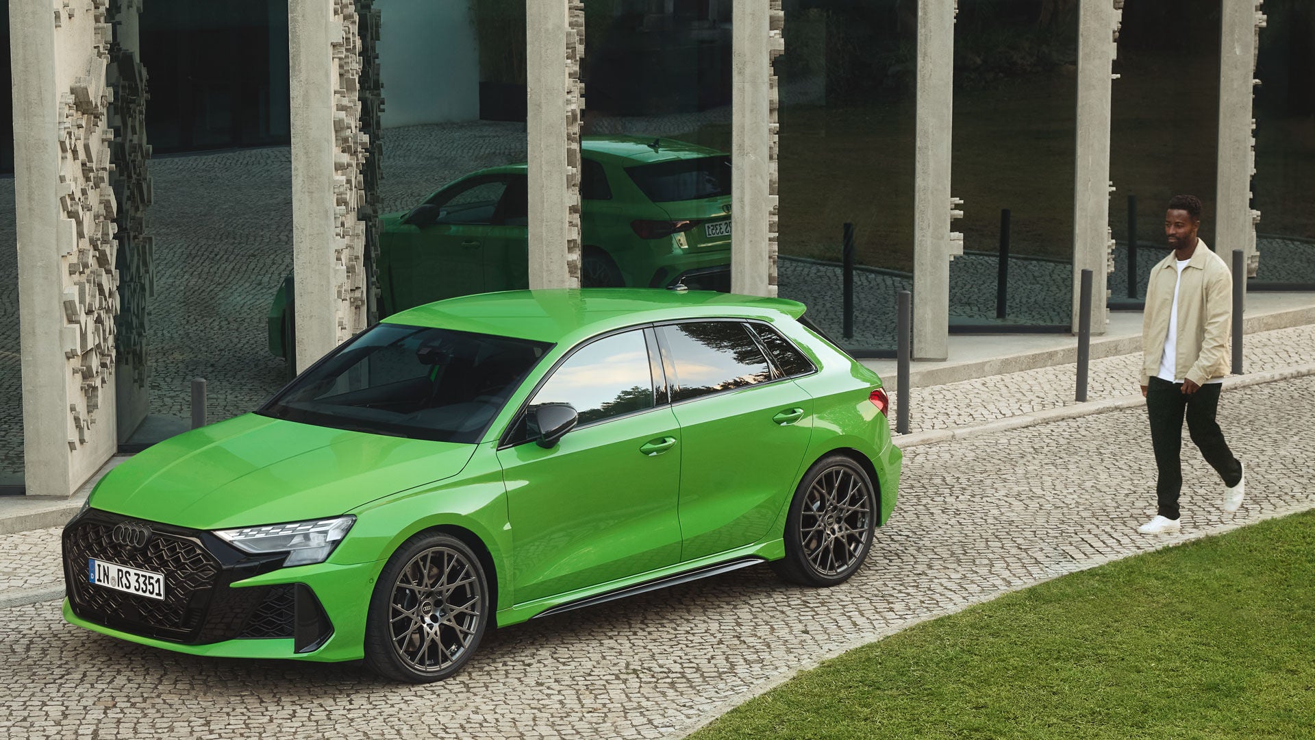 Grüner Audi RS 3 Sportback vor Glasfassade geparkt, ein Mann in heller Jacke nähert sich auf gepflastertem Weg dem Fahrzeug.