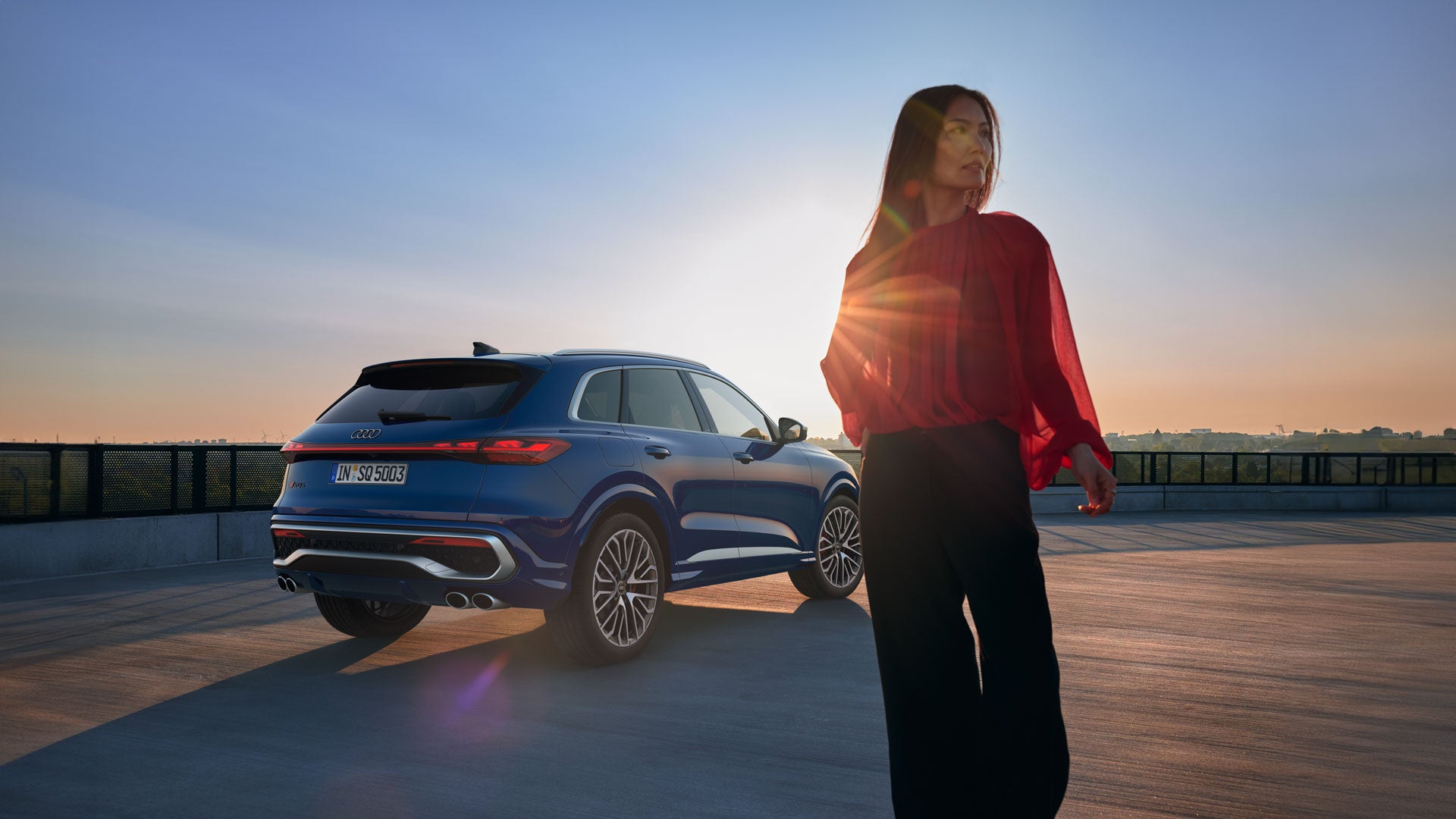 Frau im roten Oberteil steht neben einem blauen Audi SUV bei Sonnenuntergang auf einem Parkdeck mit weitem Ausblick.