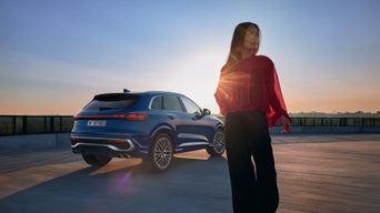 Frau im roten Oberteil steht neben einem blauen Audi SUV bei Sonnenuntergang auf einem Parkdeck mit weitem Ausblick.