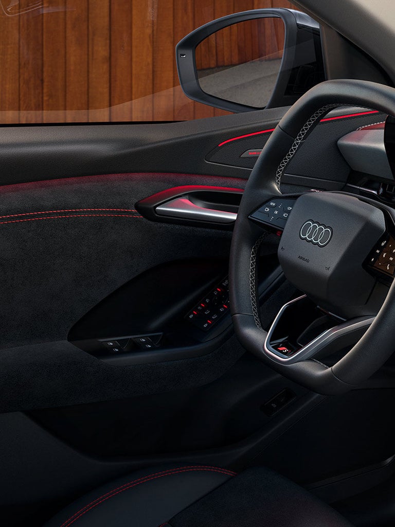 Nahaufnahme des Lenkrads mit Audi Logo, schwarzer Türverkleidung mit roten Nähten und Türbedienelementen.