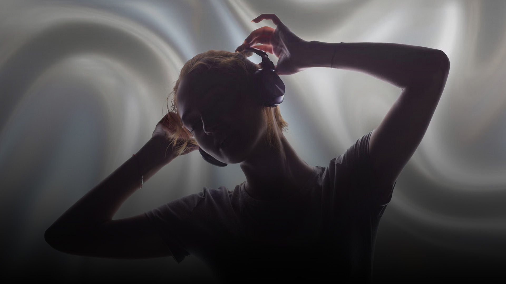 Person mit Kopfhörern im Profil hebt die Hände an den Kopfhörer, vor hellem, weich verschwommenem Hintergrund.