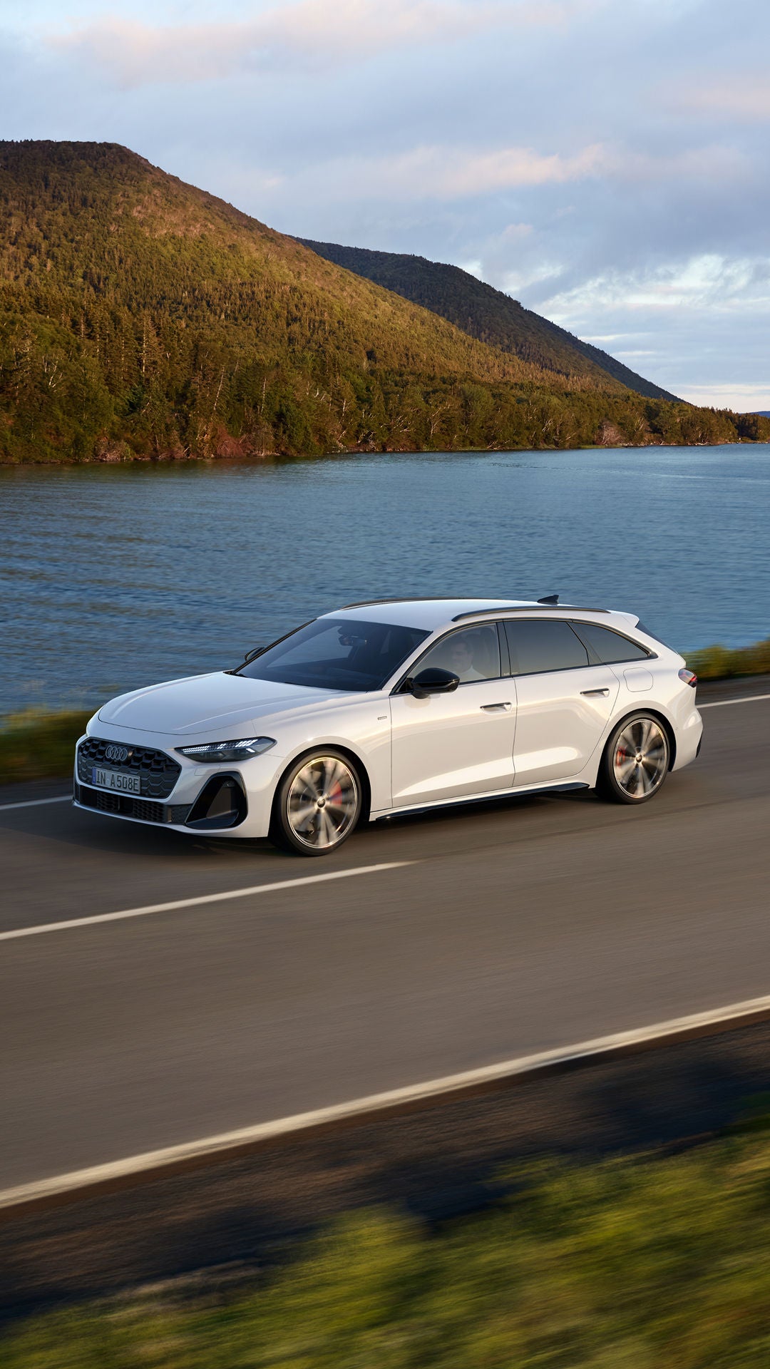 A sleek white Audi A5 Avant e-hybrid drives along a scenic coastal road with calm waters and greenery in the background.