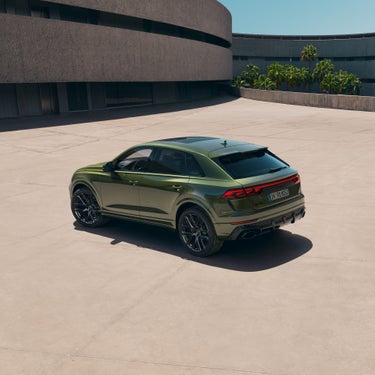 Ein grüner Audi RS Q8 SUV aus schräger Heckansicht auf heller Betonfläche vor moderner Architektur und Palmen im Hintergrund.