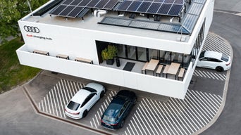 Audi charging hub mit Solarpaneelen auf dem Dach, drei Elektroautos auf markierten Ladeplätzen und Sitzbereich im Obergeschoss.