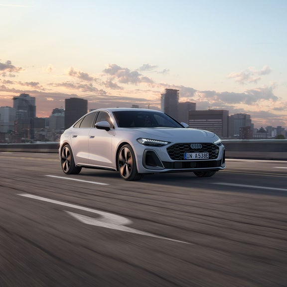 Silver car driving on an open highway at sunset, with a city skyline in the background.
