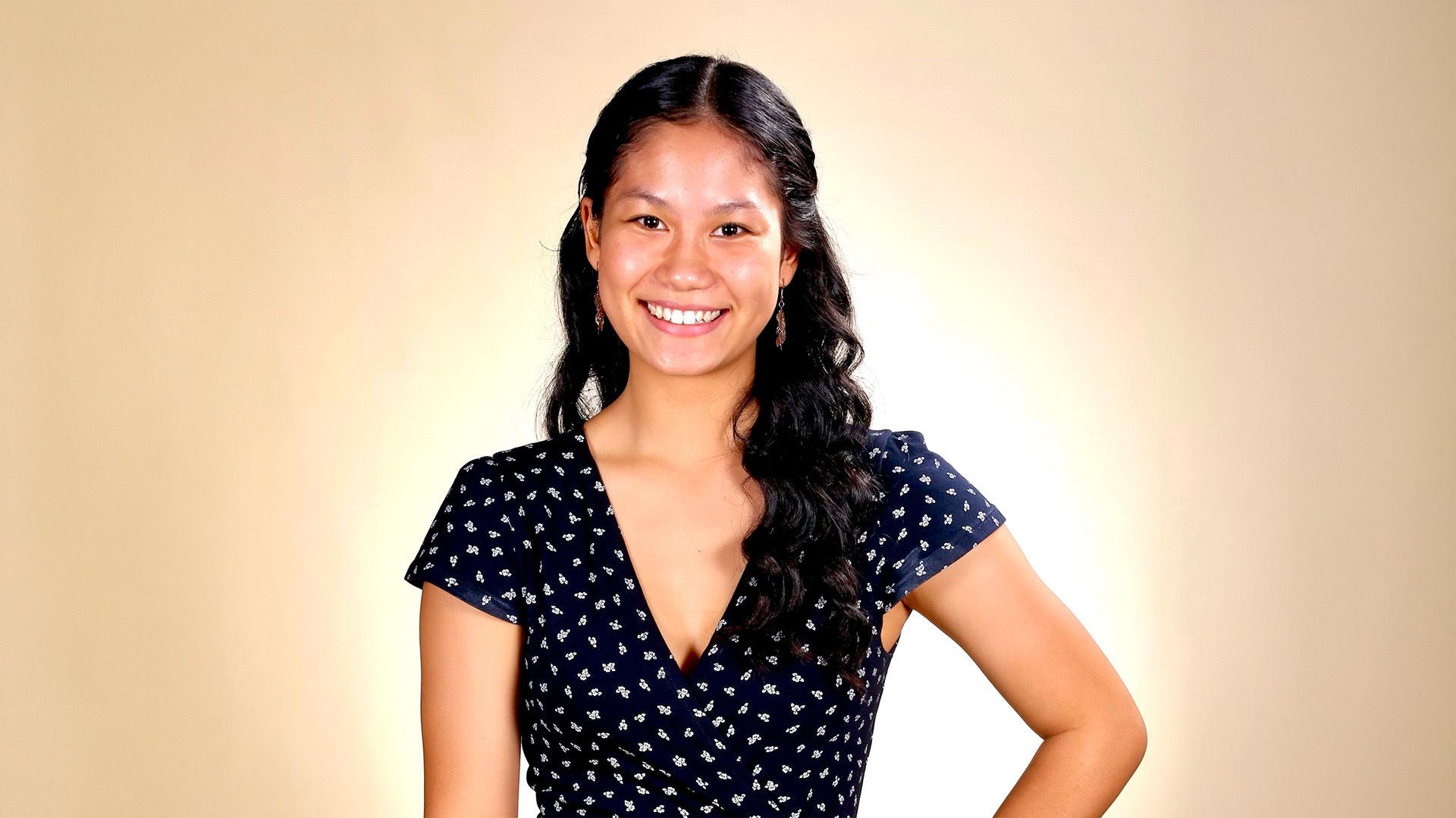 Frau mit langen dunklen Haaren im dunkelblauen Kleid mit Muster, lächelnd, vor beigem Hintergrund.