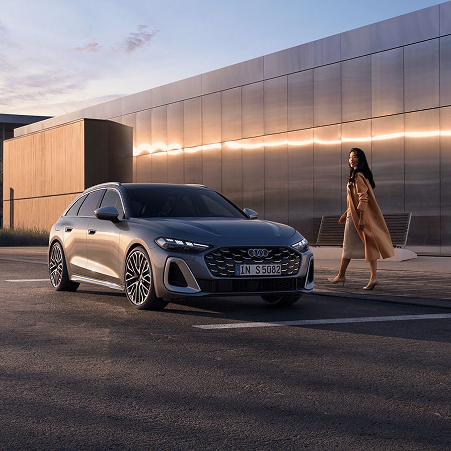 A sleek silver Audi parked in front of a modern building during twilight, with a woman elegantly walking nearby.