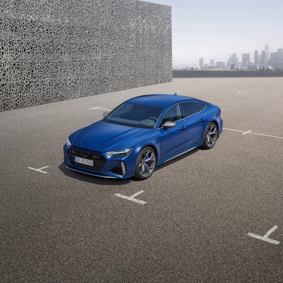 Blue sedan parked on concrete with an intricate wall design and city skyline in the background.