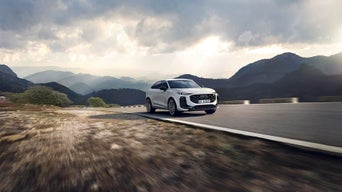 Ein weißer Audi fährt auf Bergstraße, dunkle Wolken und Berglandschaft im Hintergrund.
