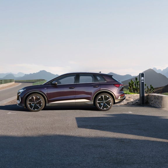 Electric SUV in deep purple, parked at a charging station by a winding road. Mountains and clear sky are visible in the background.