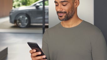 Mann im grauen Shirt blickt lächelnd auf Smartphone, unscharfer Audi im Hintergrund durch Glasfront sichtbar.