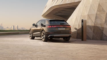 The Q8 SUV e-hybrid is parked in an angled rear view next to a futuristic building, with the skyline of a major city in the background. The charging plug of a charging station, visible behind the vehicle, is connected to the Q8 for charging.