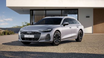 Une A6 Avant argentée est garée sur un terrain gravelé devant une maison individuelle moderne.