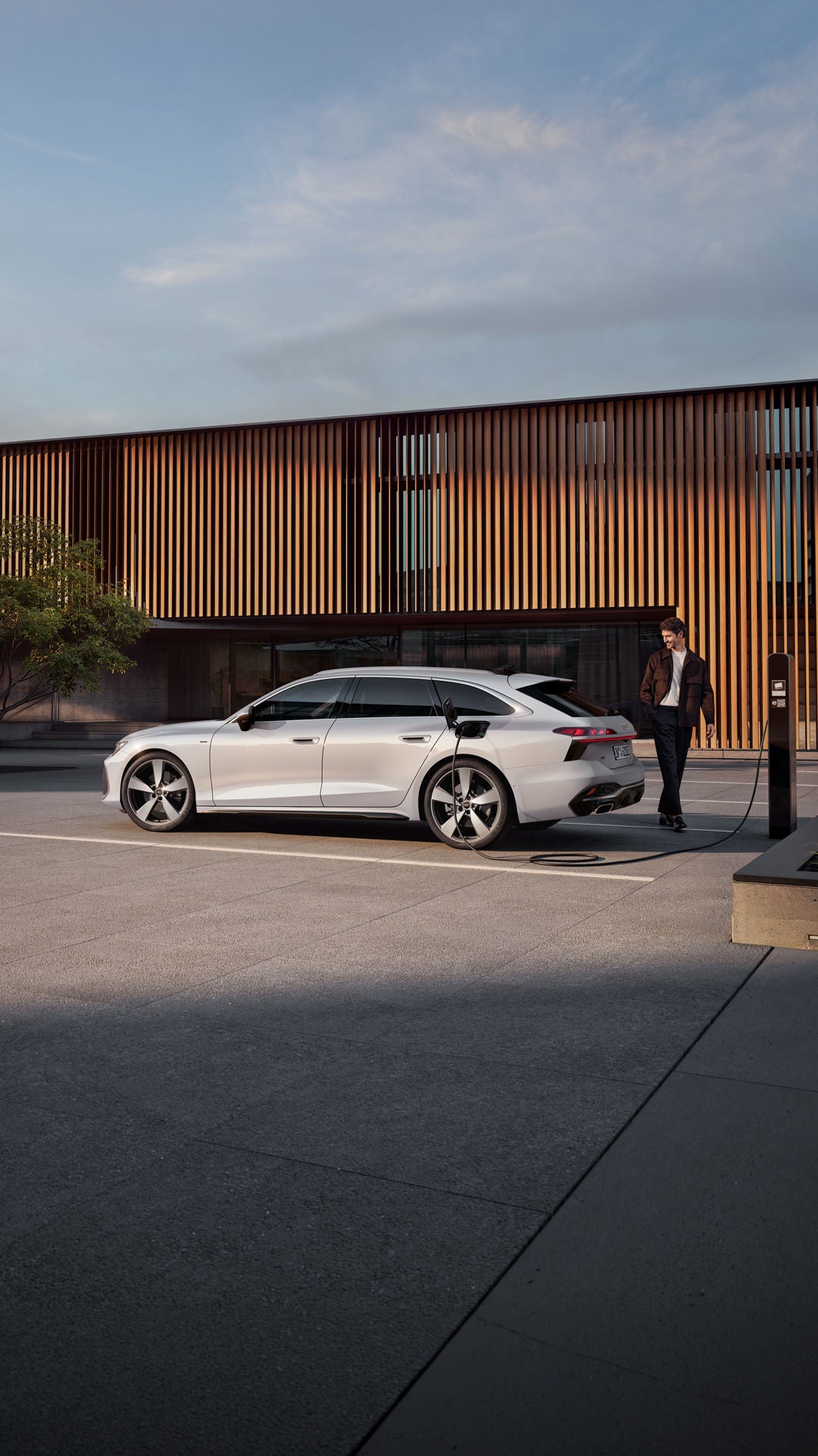 Der A6 Avant e-hybrid steht seitlich vor dem Eingangsbereich eines großen Gebäudes und wird über ein Stromkabel geladen. Am Heck des Fahrzeugs steht ein Mann mittleren Alters.