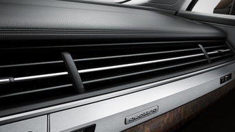 Nahaufnahme eines Lüftungsgitters im Innenraum eines Audi-Fahrzeugs mit „quattro"-Schriftzug auf einer metallischen Blende. Die Materialien wirken hochwertig, mit einer Kombination aus Aluminium, Holzdekor und Leder.