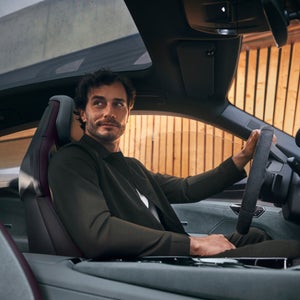 A man in a dark suit sits confidently in a car, holding the steering wheel. Wooden walls and a sunroof filter warm light into the sleek interior.