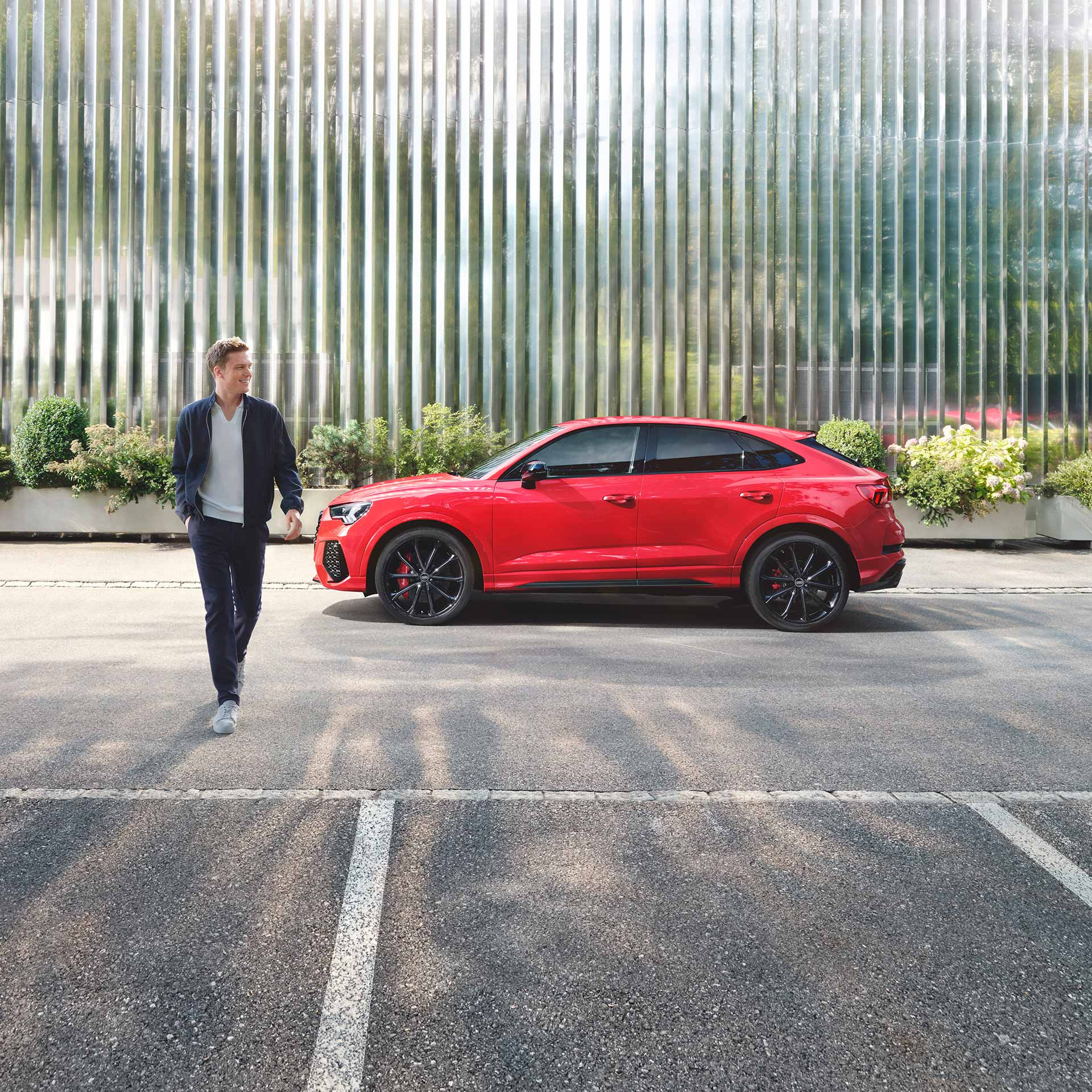 A person walking past a red car parked beside a metal corrugated fence and greenery.