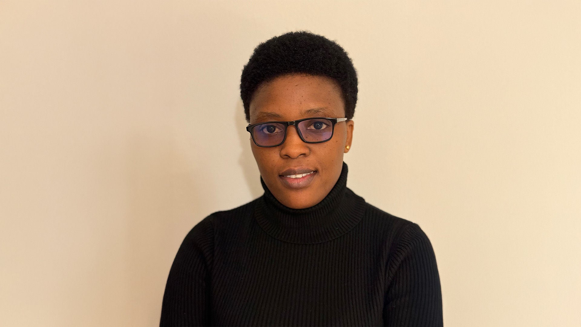 Porträt einer Frau mit kurzen Haaren und Brille im schwarzen Rollkragenpullover, neutraler Gesichtsausdruck, beiger Hintergrund.