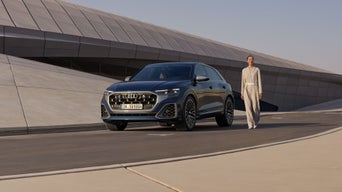 Der SQ8 SUV vor einem futuristischen Gebäude mit geschwungener Form parkend, daneben eine modisch gekleidete junge Frau.