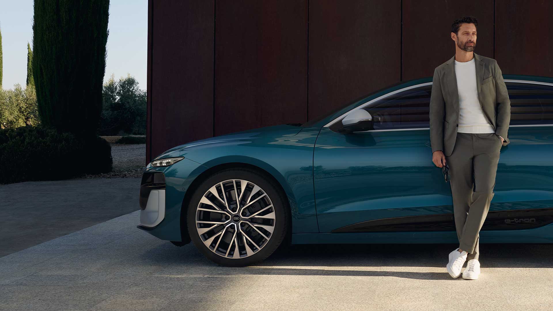 Ein Mann lehnt lässig an einem grünen Audi S6 Sportback e-tron, der seitlich vor einer dunklen Wand geparkt ist.
