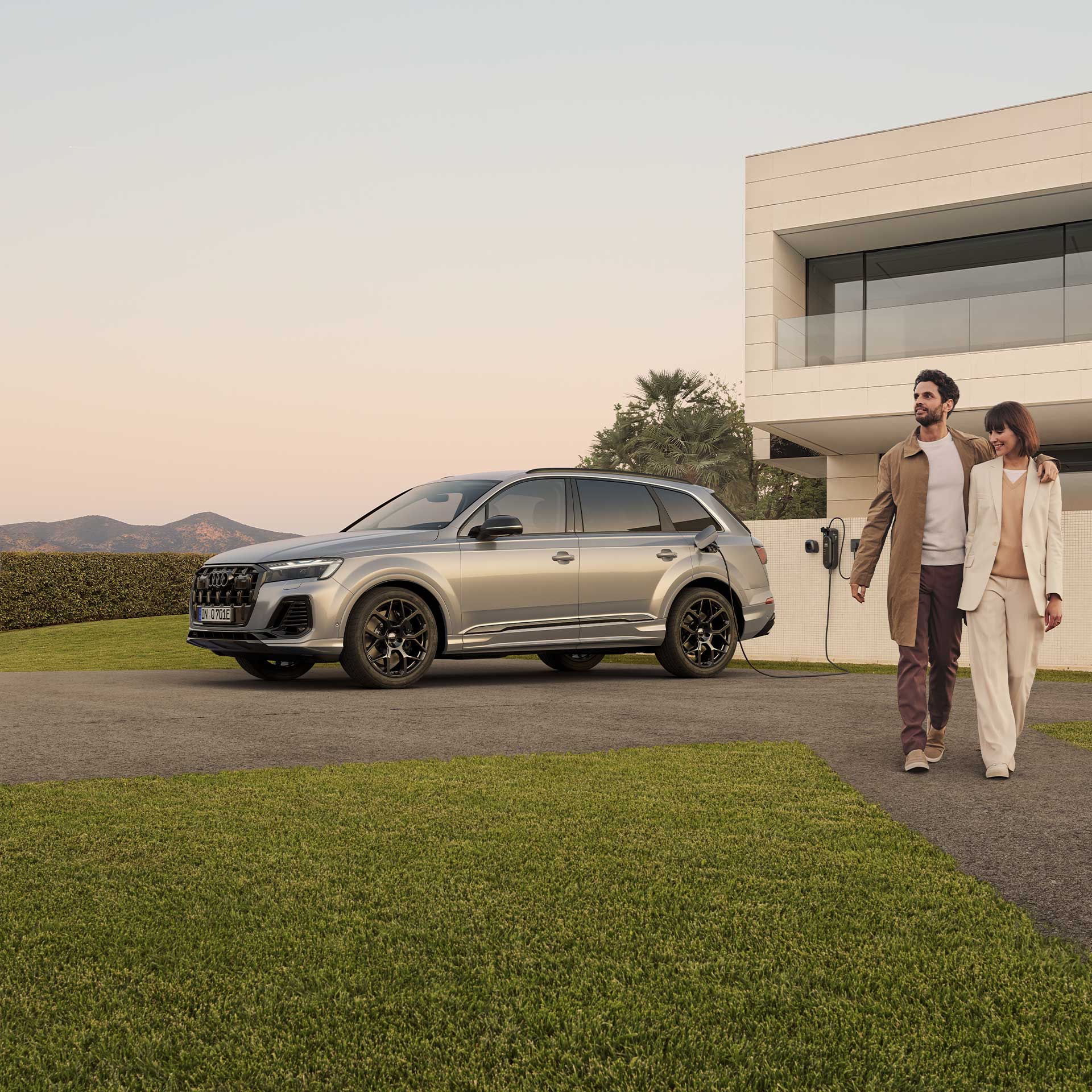 Ein silberner Audi Q7 SUV TFSI e lädt an Wallbox vor modernem Haus, ein lächelndes Paar im Businessoutfit läuft Arm in Arm rechts am Auto vorbei.
