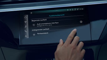Person im Audi-Innenraum bedient Touchscreen mit Fahrzeugfunktionen, digitales Cockpit und Klimaeinstellungen sichtbar.