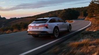 De Audi A6 allroad e-hybrid, schuin van achteren gezien terwijl hij op volle snelheid langs een bergweg rijdt. Op de achtergrond is een bosrijk gebied te zien.