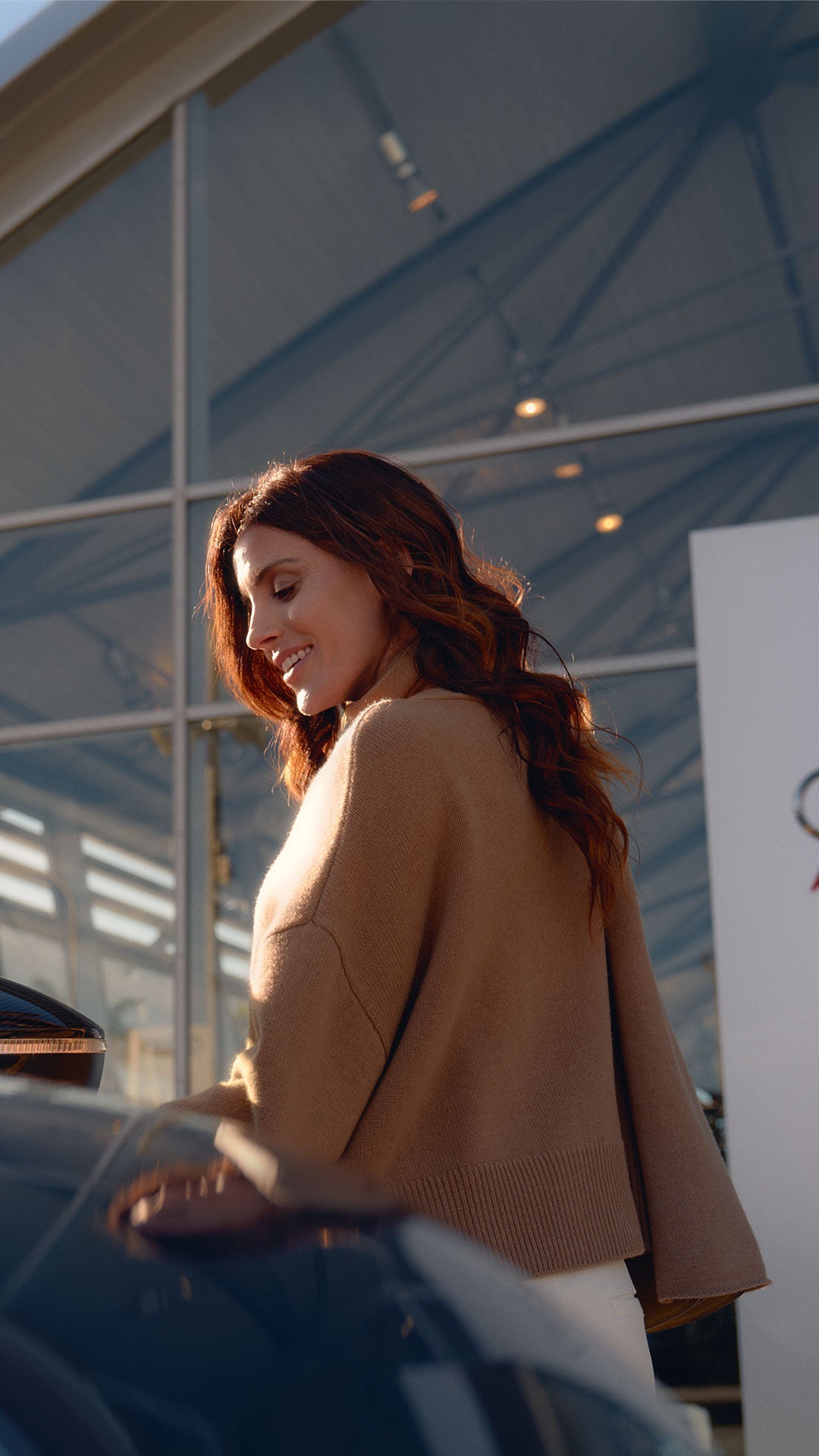 Une femme rousse vêtue d'un manteau marron se tient devant un concessionnaire Audi et monte dans une voiture.