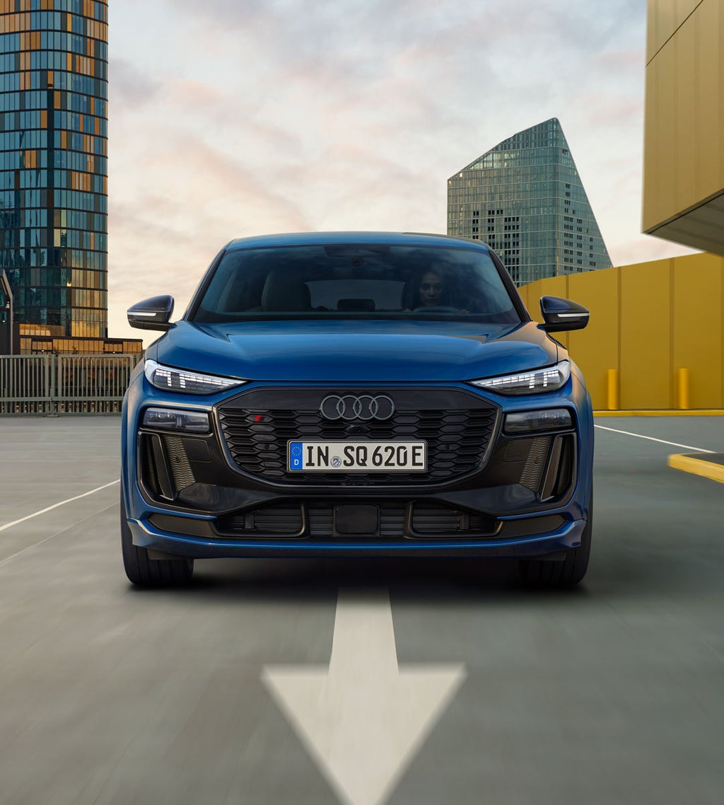 Frontaufnahme des SQ6 Sportback e-tron, welcher über eine Straße in der Stadt fährt. Im Hintergrund sind gläserne Hochhäuser zu sehen.
