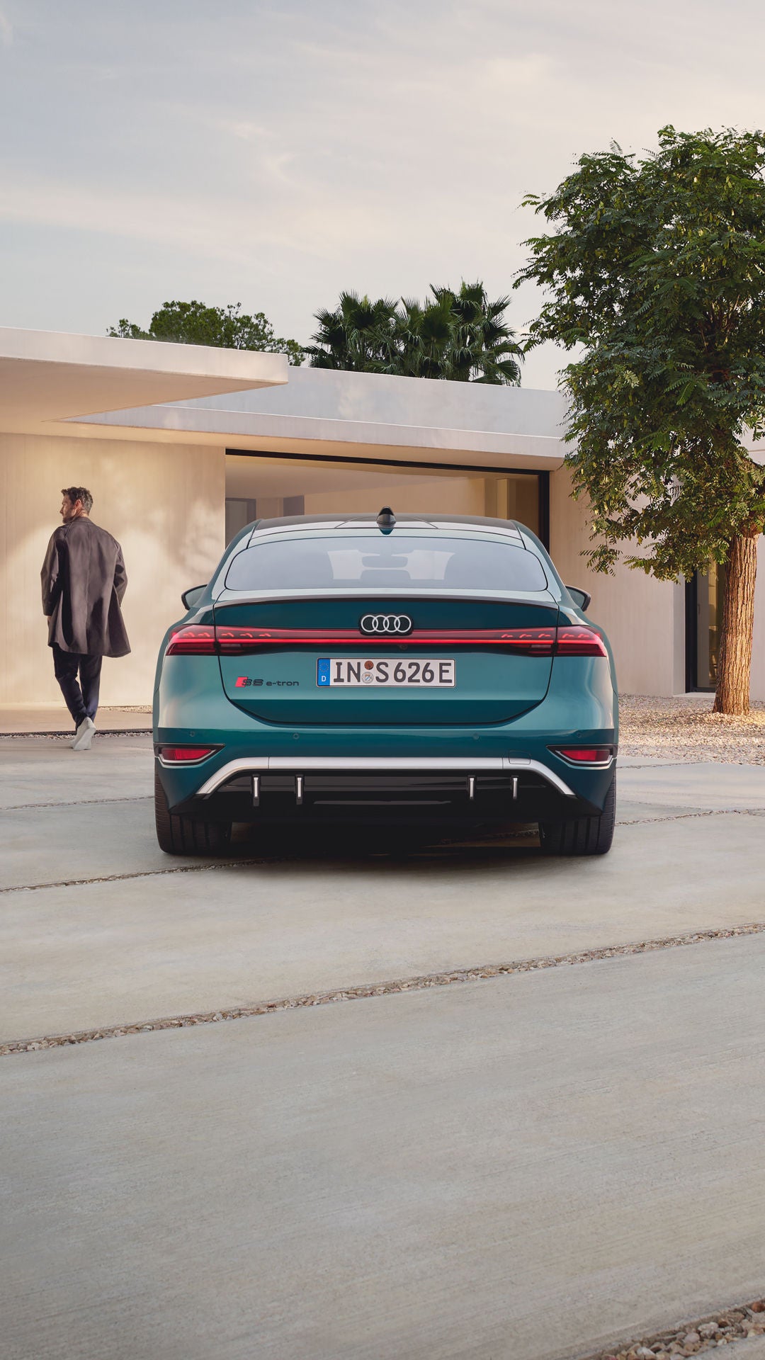 Der S6 Sportback e-tron im Heckprofil. Das Fahrzeug ist dem Eingangsbereich eines modernen Gebäudes zugewandt. Vor dem Auto befindet sich ein junger Mann, der auf das Gebäude zuläuft.