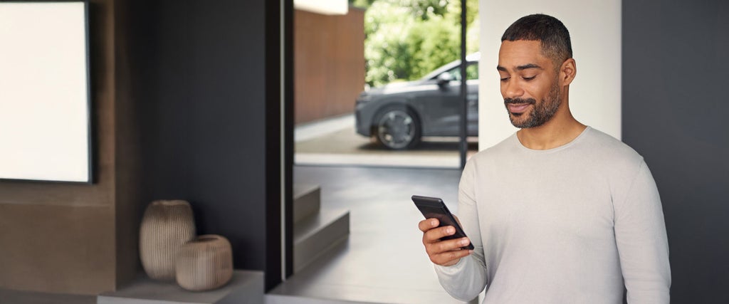 Zwei Männer stehen in einer Autowerkstatt neben einem geöffneten Auto mit sichtbaren orangefarbenen Hochvoltkabeln. Der linke Mann trägt ein schwarzes Polohemd mit Audi-Logo und hält ein Tablet, auf das beide schauen. Im Hintergrund sind zwei silberne Audi-Fahrzeuge zu sehen.